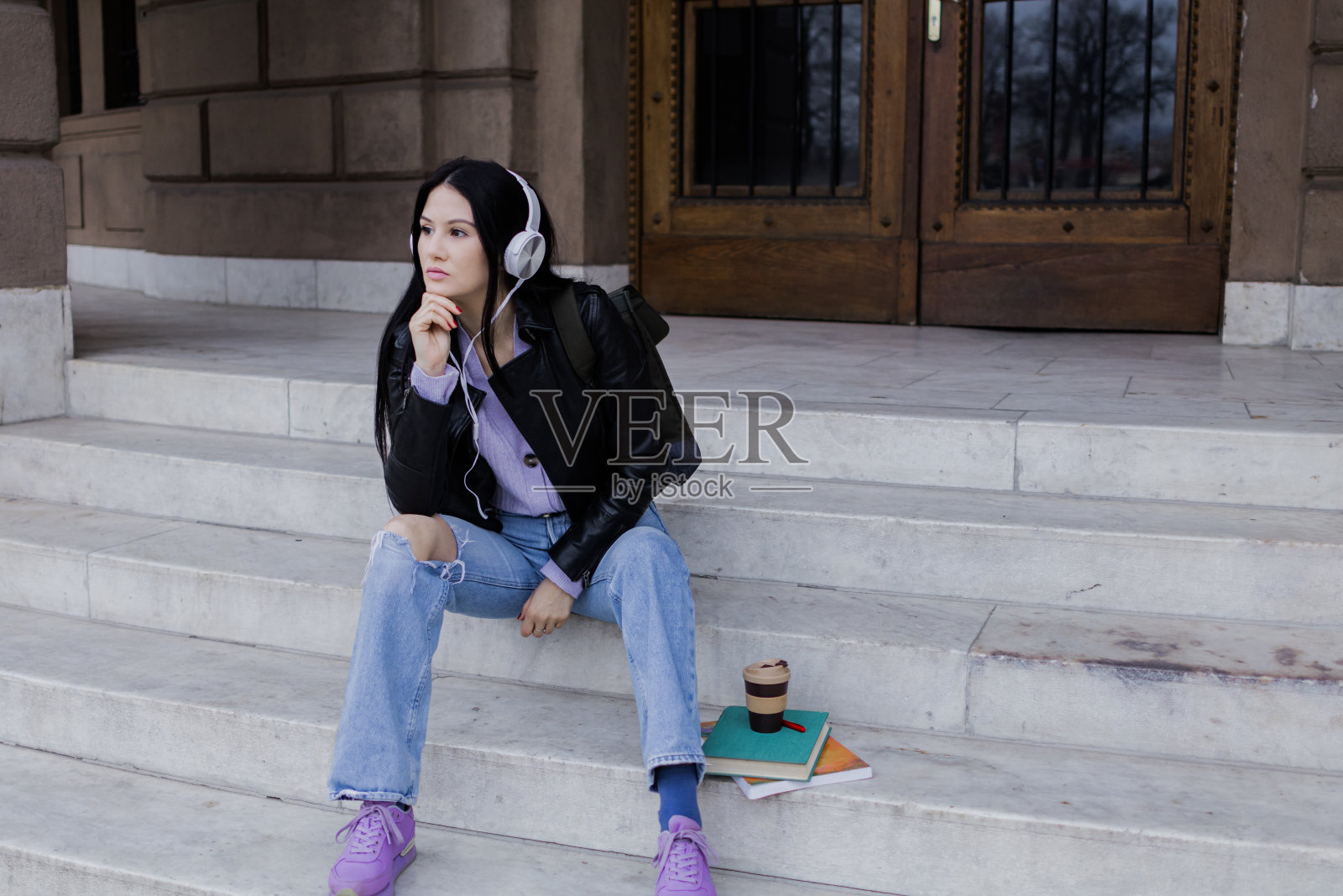 年轻漂亮的女大学生背着双肩包坐在大学楼梯上。她戴着耳机，穿着皮夹克，等待着大学课程的开始。照片摄影图片
