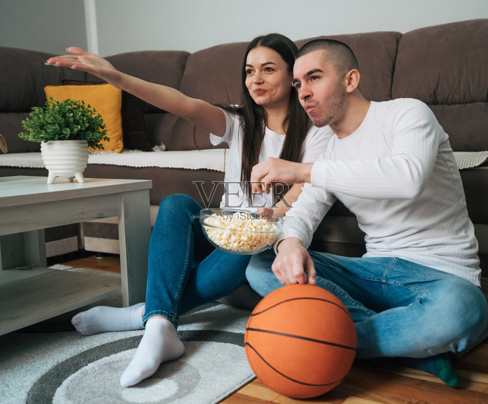 一对年轻夫妇坐在家里的地板上看篮球比赛。照片摄影图片