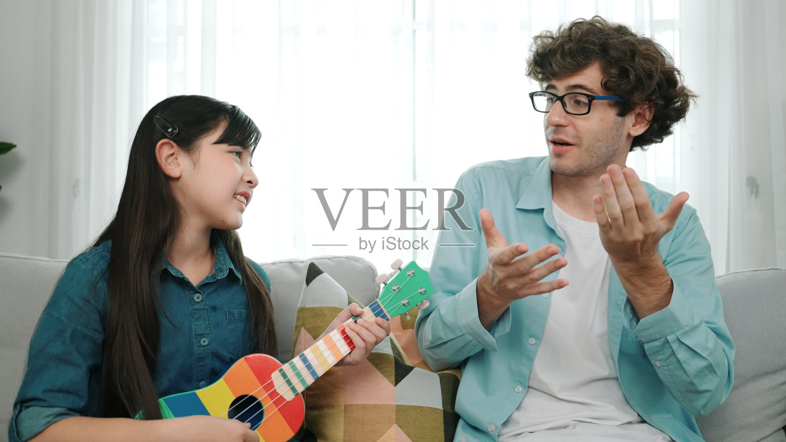 父亲看着聪明的女儿热情地弹奏尤克里里的背影。教育学。照片摄影图片