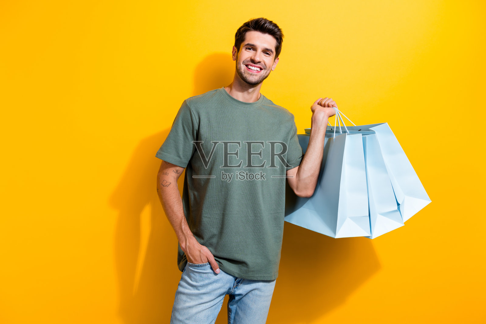 可爱的家伙的肖像，鬃毛，穿着时尚的t恤，拿着购物袋，手臂放在口袋里，鲜艳的黄色背景孤立照片摄影图片