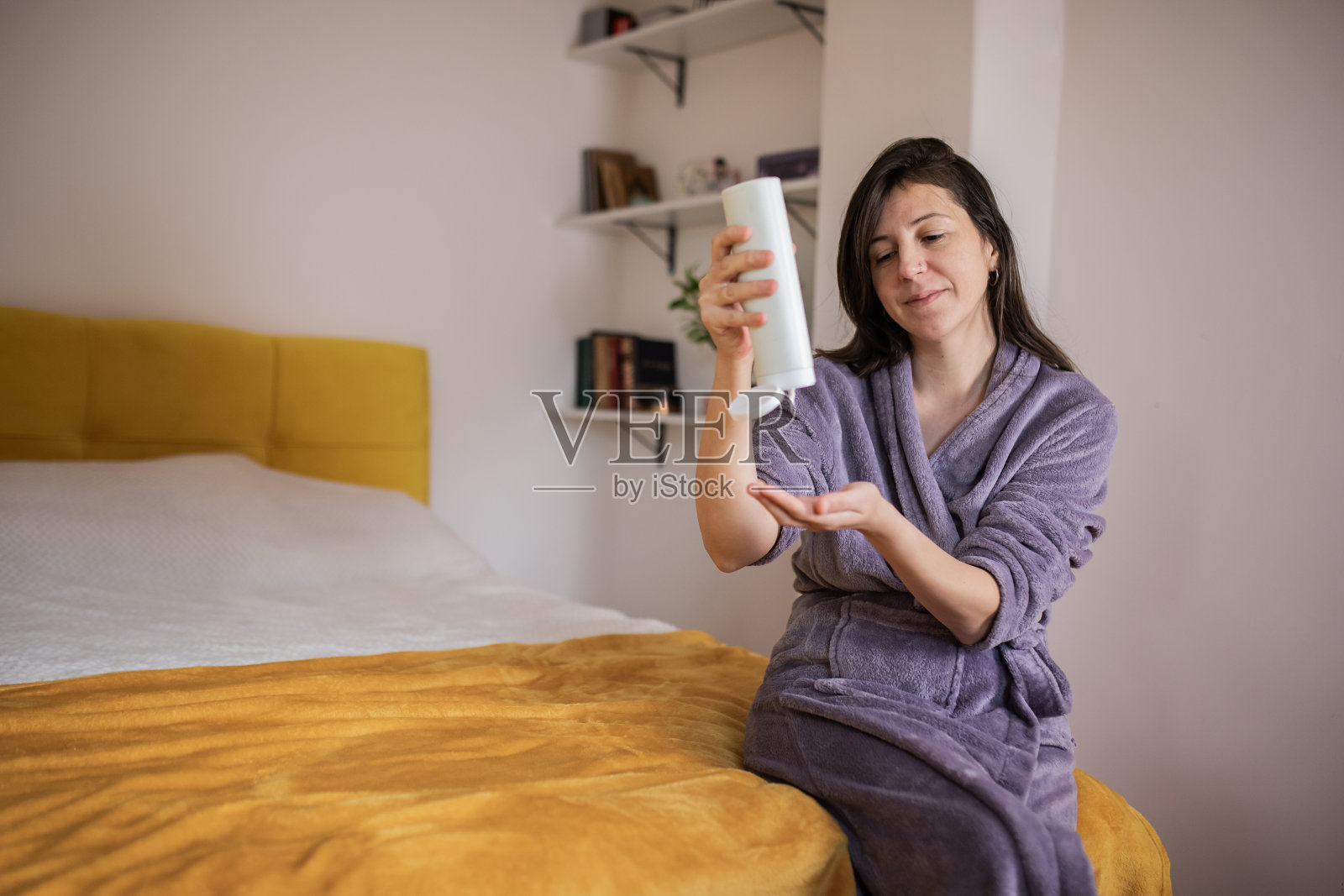 年轻漂亮的女人坐在床上，洗澡后在身上涂乳液。照片摄影图片