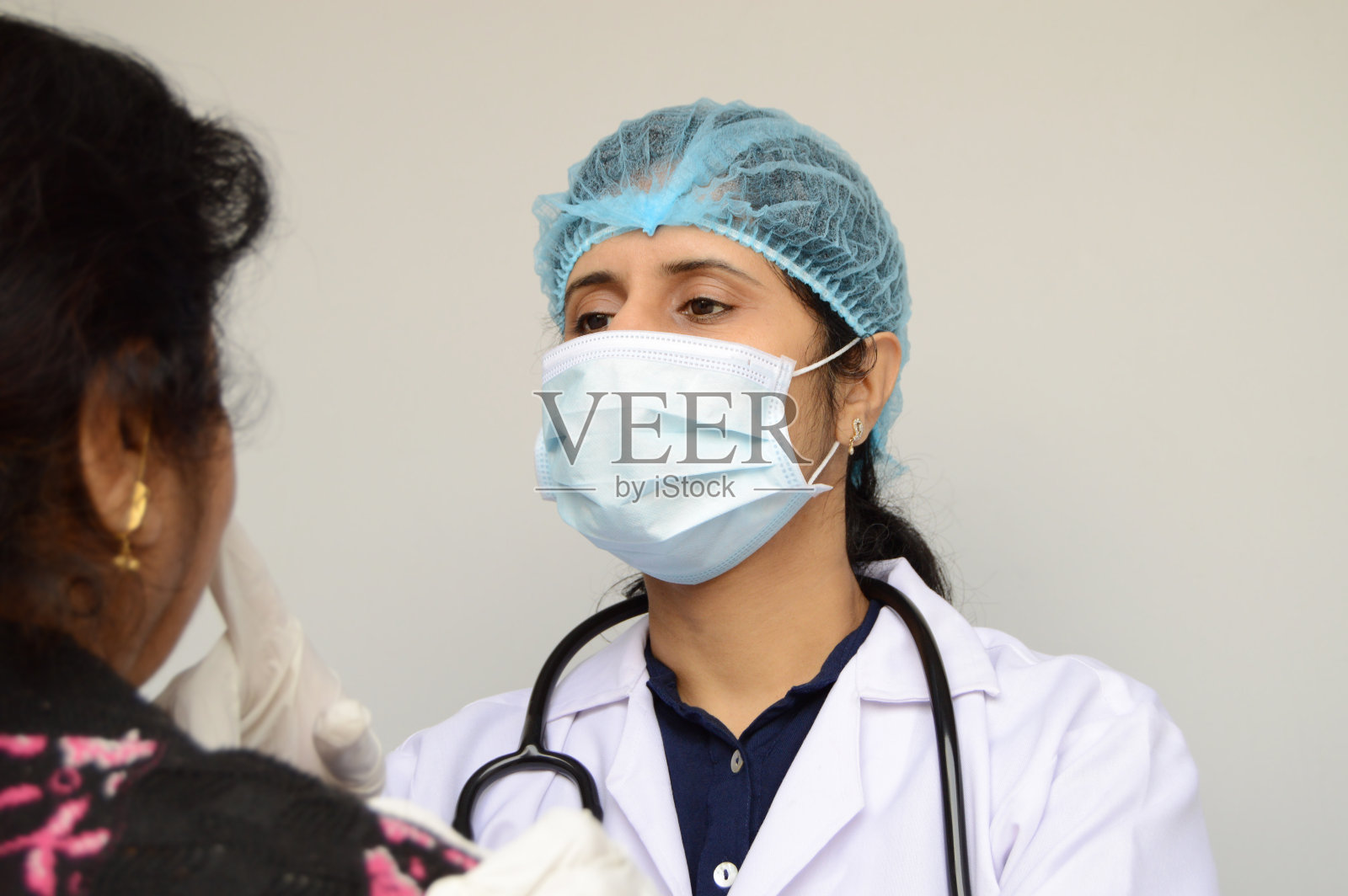 在灰色的水平背景上，戴着外科口罩，戴着外科帽，穿着白色大褂，脖子上挂着听诊器，用手指看着和触摸病人的脸，专心地检查病人照片摄影图片
