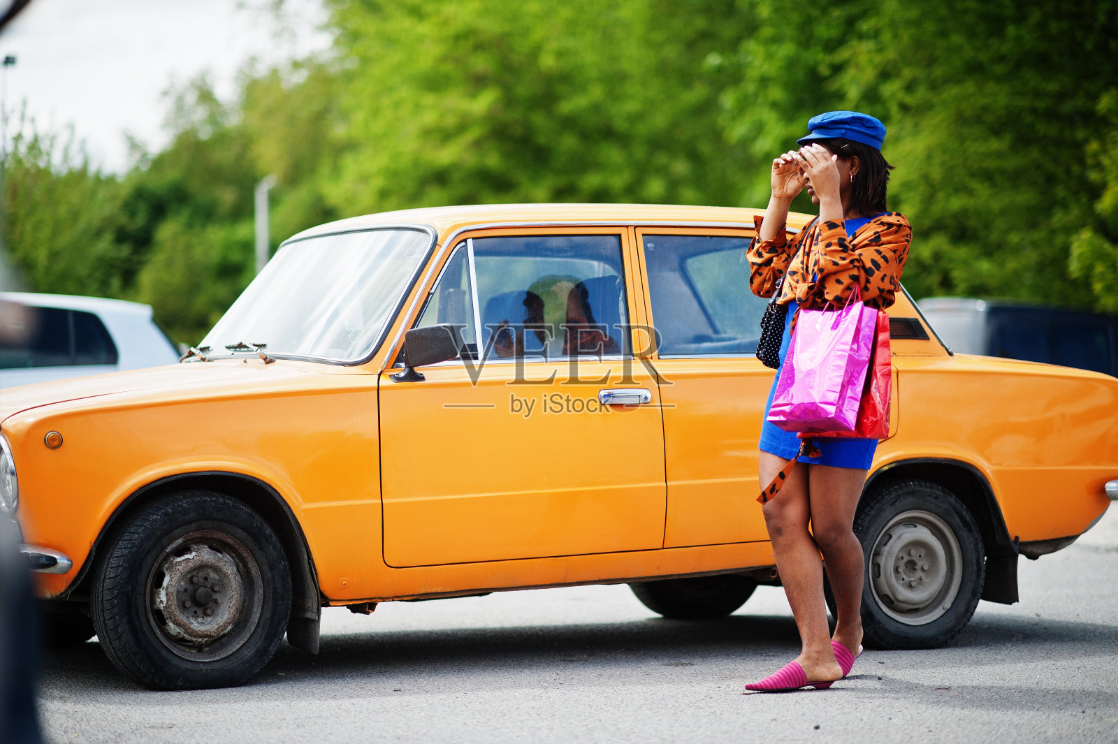 漂亮的非裔美国女士拎着购物袋站在橙色经典复古车旁。照片摄影图片