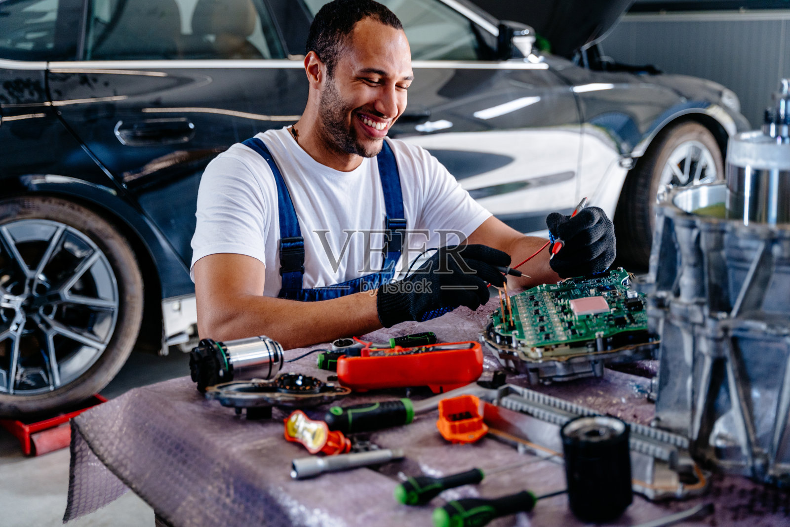 阿拉伯男性汽车修理工用电压表检查从汽车上拆下来修理的汽车发动机。现代电动汽车的电控单元(ECU)。照片摄影图片