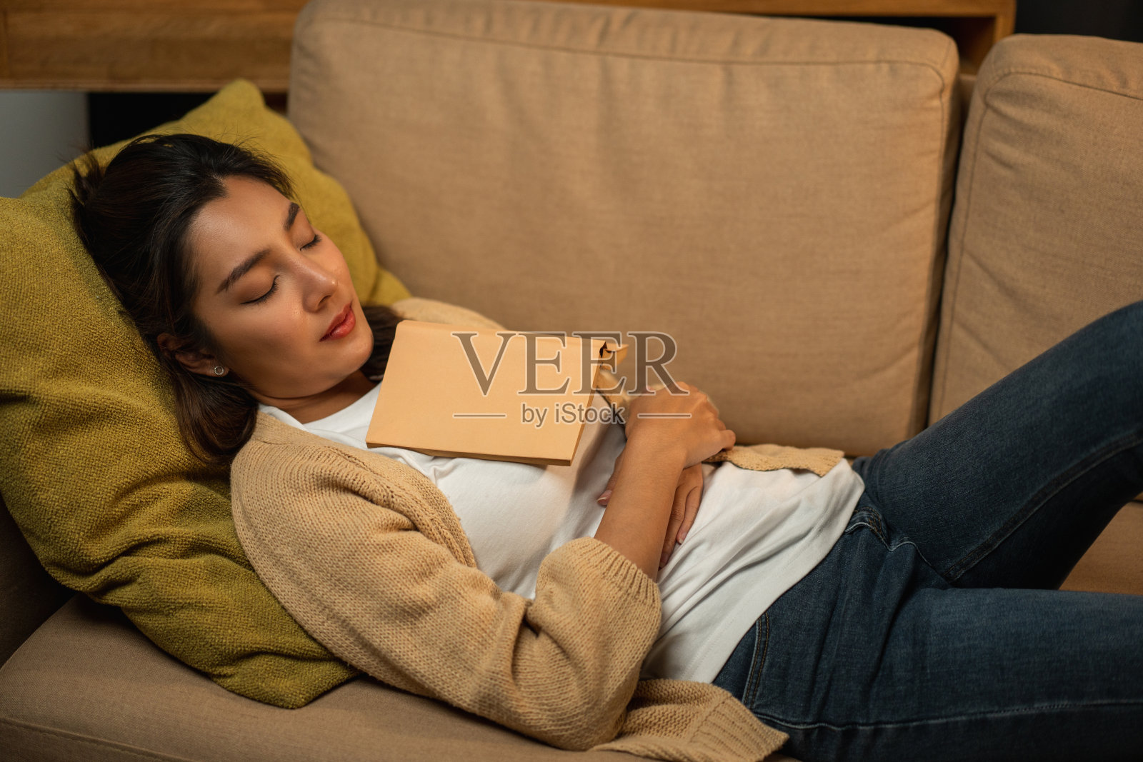 睡觉的亚洲美女躺在舒适的沙发上，晚上在客厅里温暖的灯光下看书。人们女性看书直到入睡，业余时间呆在家里放松照片摄影图片