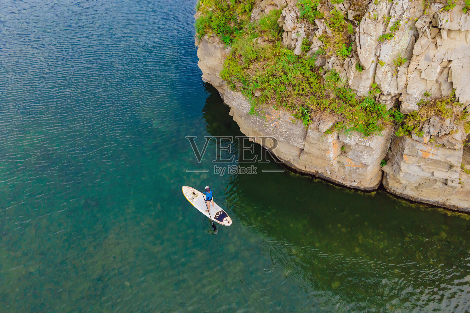 年轻女子站着在海里划桨玩得很开心。喂，红发女孩在岩石附近的桨板上训练照片摄影图片
