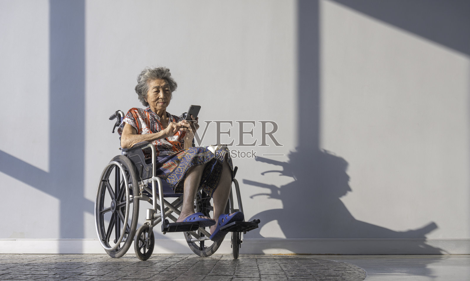 亚洲精神健康的老年妇女在医院内等待年度体检时坐在轮椅上玩手机。照片摄影图片