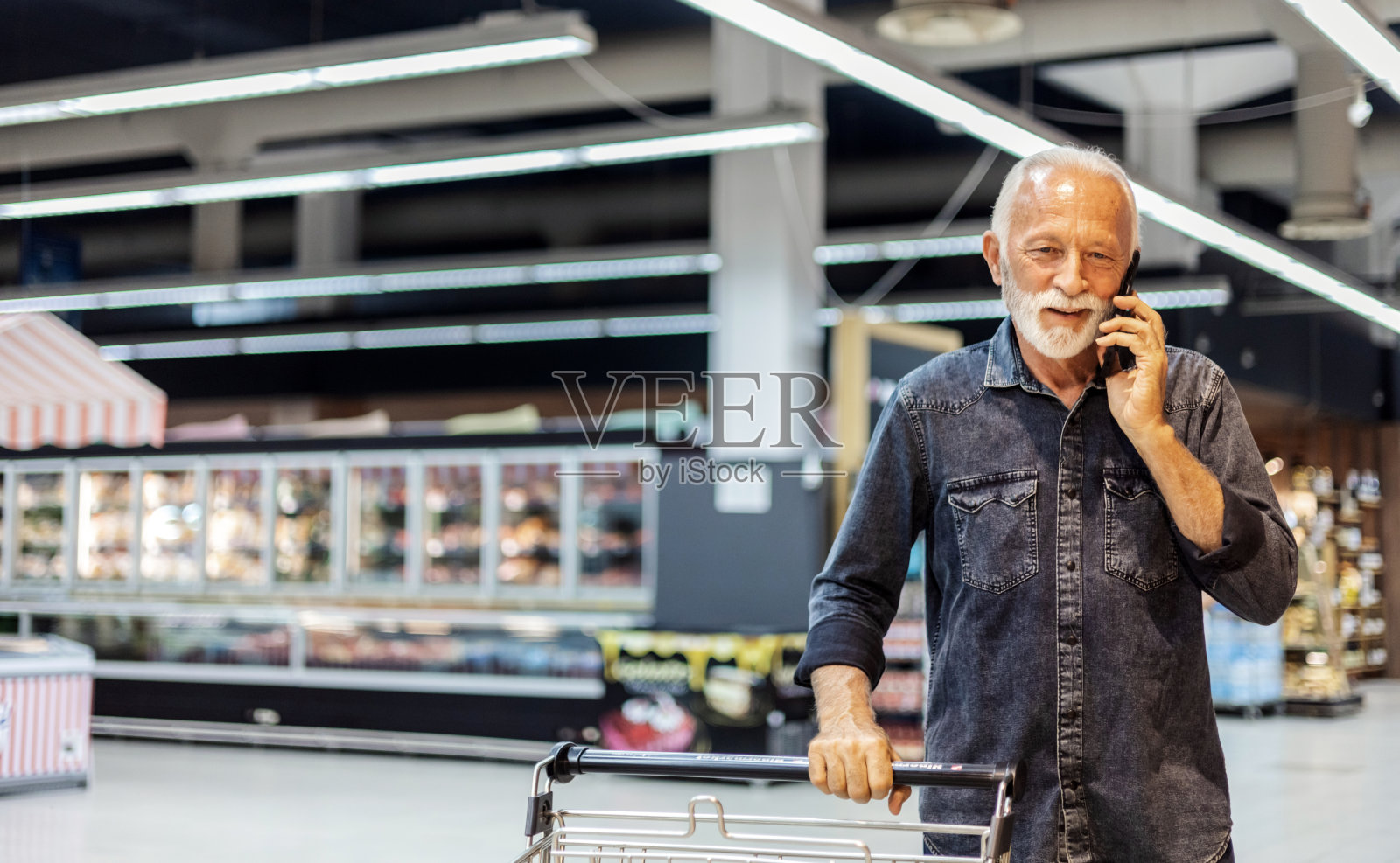一位年长的白人男子在超市购物时正在接电话。一位老人站在超市里打电话，前面放着一辆购物车。照片摄影图片