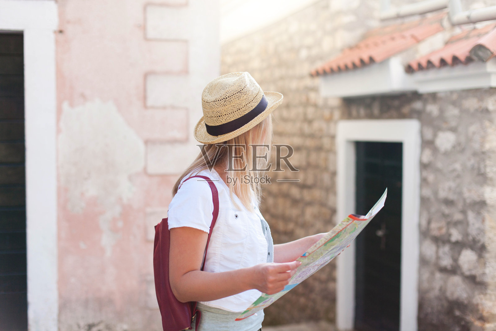 戴着帽子、背着地图和背包的女旅行者在老城区的街道上。学生旅游、暑假、女性旅游、探险、旅行、旅行的概念。女游客正在寻找公寓，住房。照片摄影图片