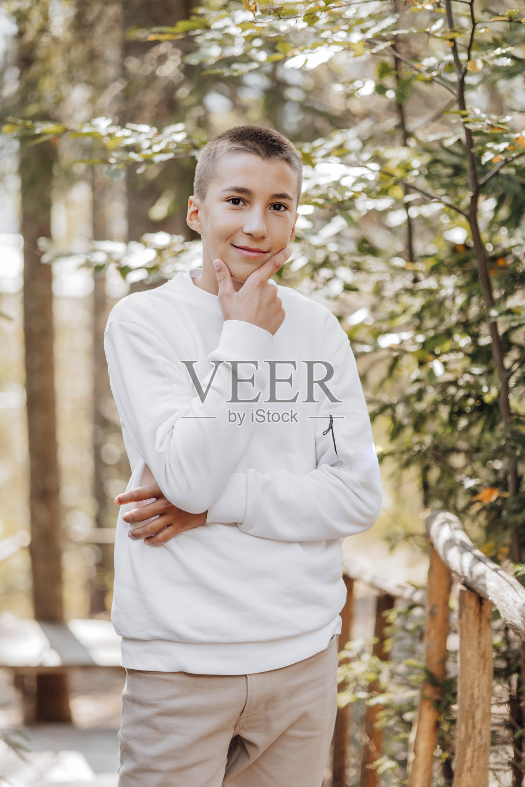 一个穿着白色毛衣和棕色裤子的少年的特写垂直肖像。夏日公园阳光下快乐微笑的少年。一个漂亮的孩子在空地上看着摄像机。照片摄影图片