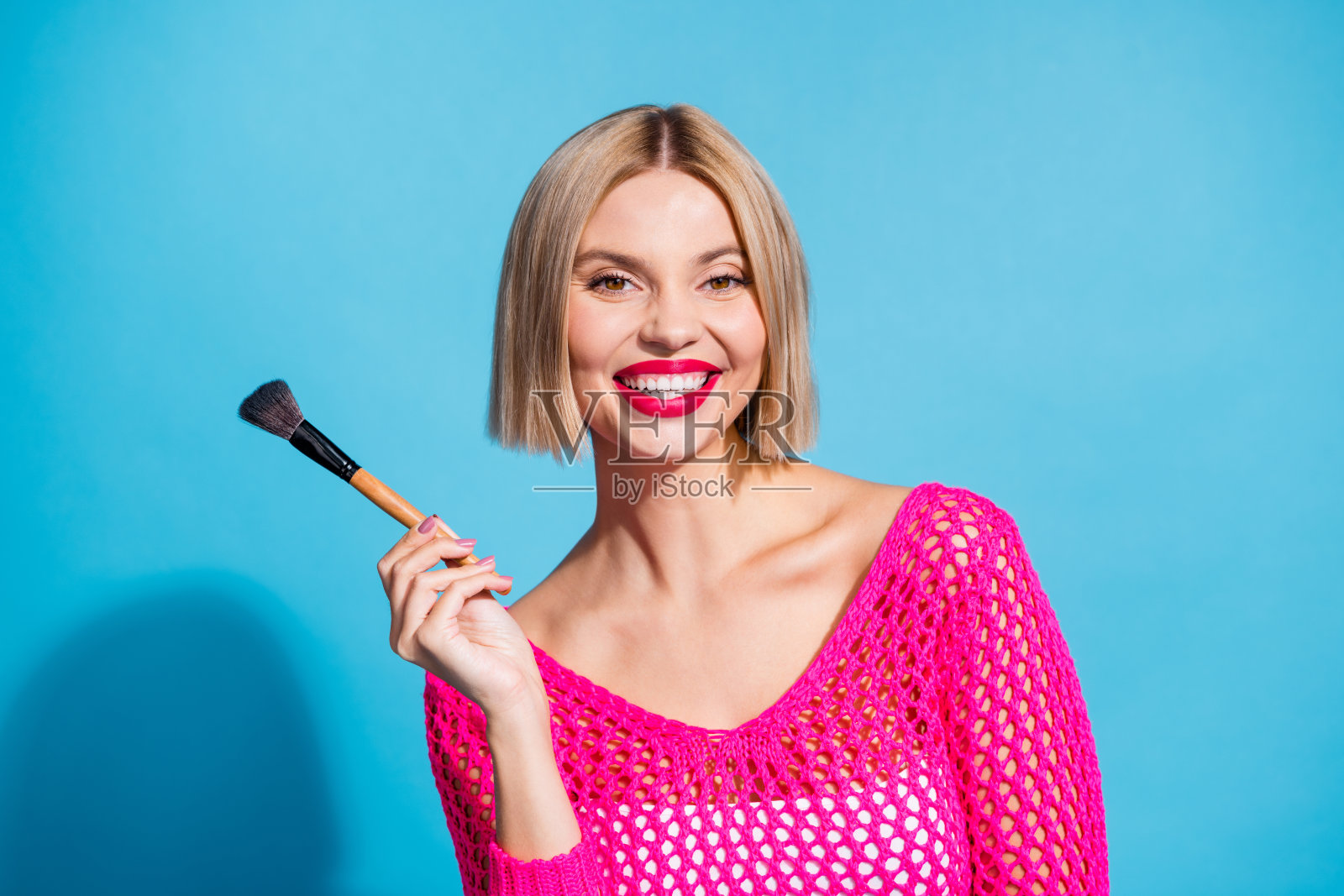漂亮的年轻女孩的照片肖像拿着画笔化妆师穿着时尚的粉红色针织服装孤立的蓝色背景照片摄影图片