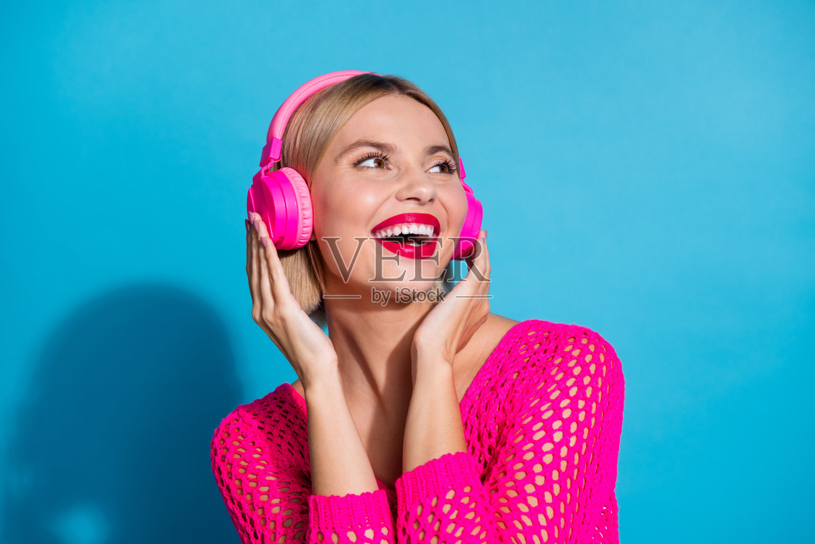 漂亮的年轻女孩的照片肖像触摸耳机看起来空的空间穿着时尚的粉红色针织服装孤立的蓝色背景照片摄影图片