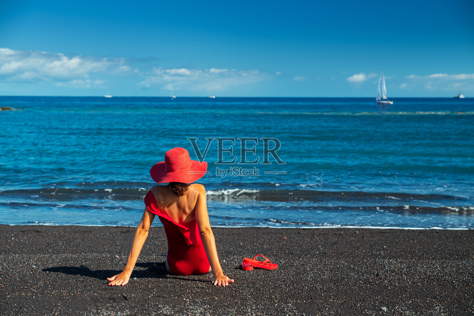 身穿红色泳衣的女子在黑色的海洋沙滩上。平静的海水和纯净的蓝天为背景。照片摄影图片