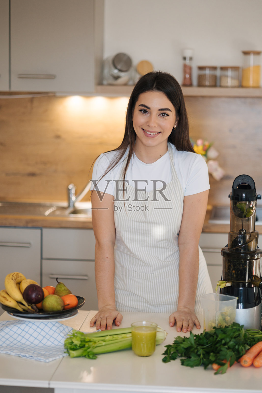 戴着围裙的女人微笑着站在厨房里。满桌的水果和蔬菜。自制排毒鸡尾酒。普通厨房的背景。香蕉、芹菜、猕猴桃等健康产品照片摄影图片