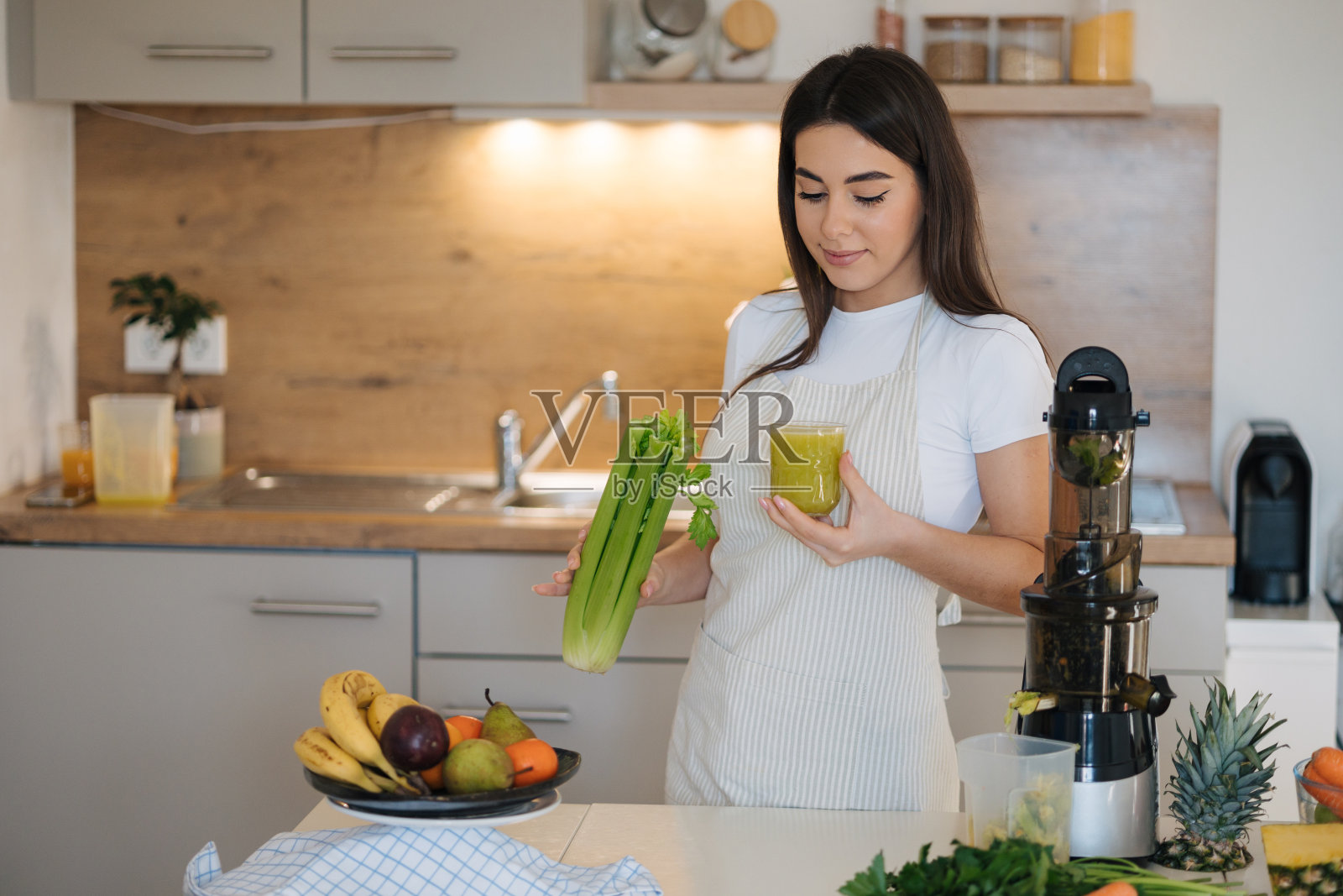 穿着围裙站在厨房里的年轻漂亮女人的画像。女子一手拿着绿色健康饮料，一手拿着芹菜。排毒饮食。自制的果汁。素食者照片摄影图片