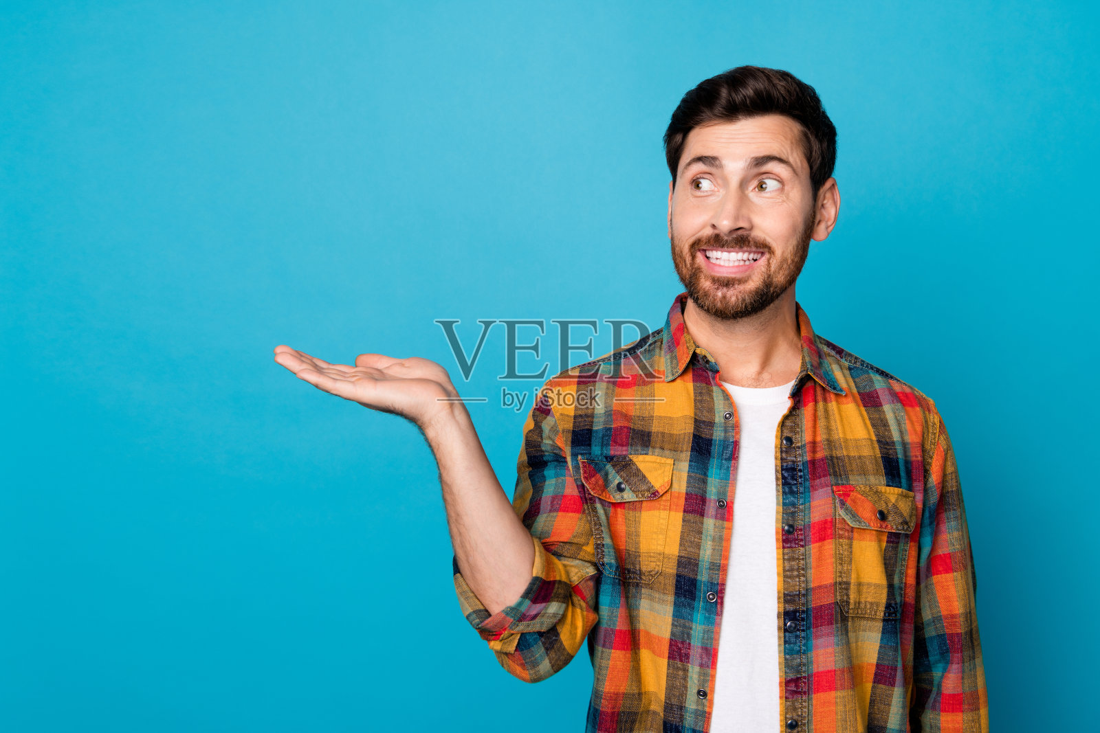 开朗的好男人的肖像，留着胡茬，穿着格子衬衫，看广告空白的手掌，孤立在蓝色背景上照片摄影图片