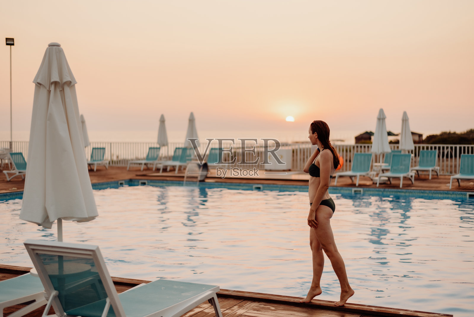 一个穿着泳衣的湿女孩站在室外游泳池的边上。夕阳下没有人照片摄影图片