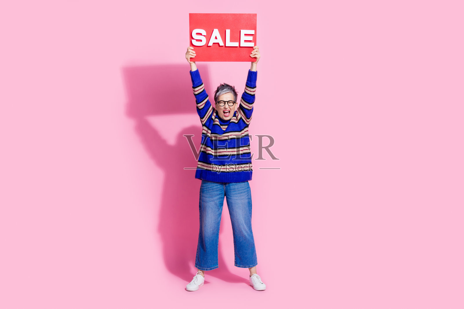 漂亮的退休女性拿着兴奋的销售盘购物的全尺寸照片，穿着时髦的蓝色条纹服装，孤立在粉红色的背景上照片摄影图片