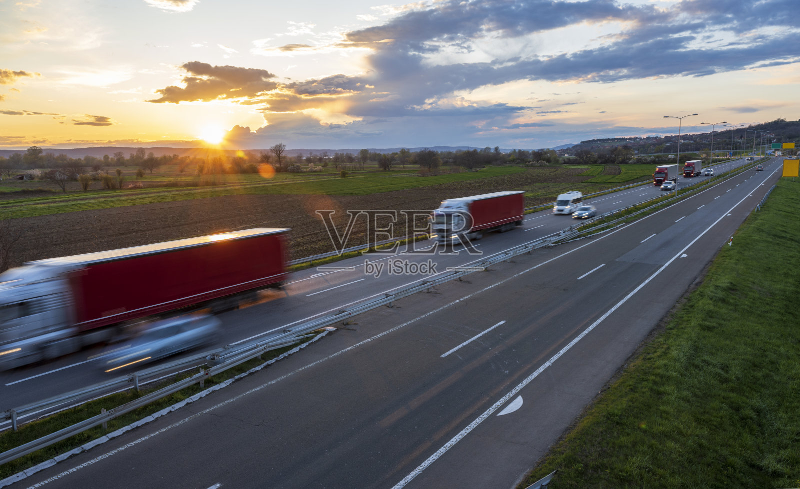 四辆红色卡车，一辆小型货车和许多汽车在乡村风景的高速公路上行驶。高速公路高速行驶。高速公路上超速行驶的景象。背景是美丽的日落。照片摄影图片
