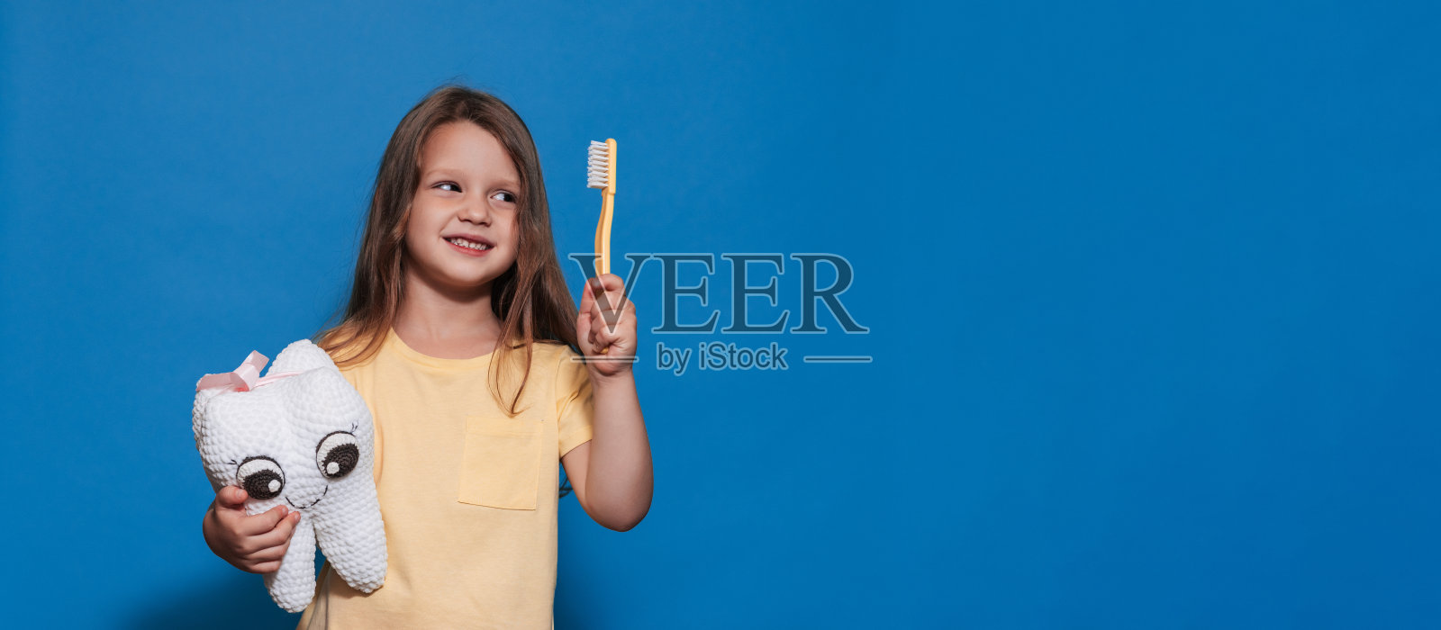 一个有着健康牙齿的微笑女孩拿着玩具牙齿和牙刷，背景是蓝色的孤立背景。放文字的地方。口腔卫生。儿科牙医。假肢。刷牙的规则。照片摄影图片