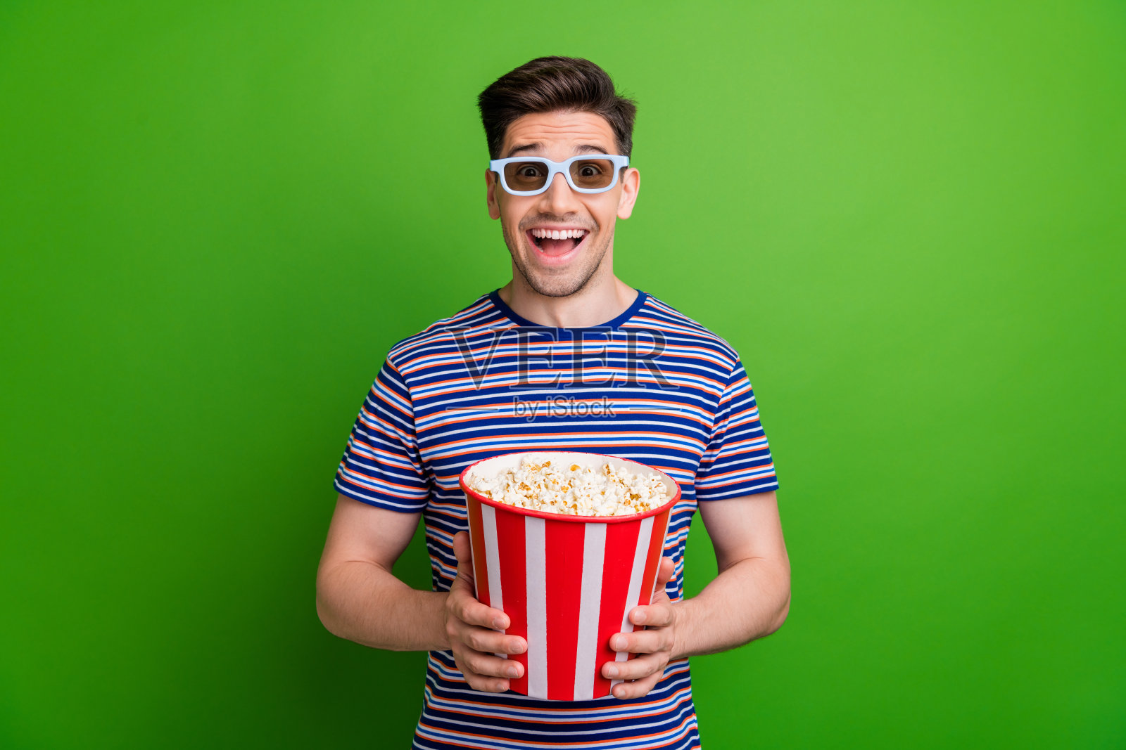 一个穿着时尚t恤，戴着3d眼镜的喜出望外的男人的肖像，在绿色的背景下，在电影院拿着爆米花照片摄影图片