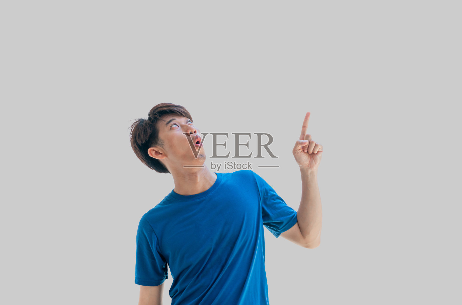 一个20多岁的亚洲年轻人，穿着蓝色t恤，开心而震惊地竖起大拇指，孤立在灰色的背景中。这个人的食指指向文本的空白区域。照片摄影图片
