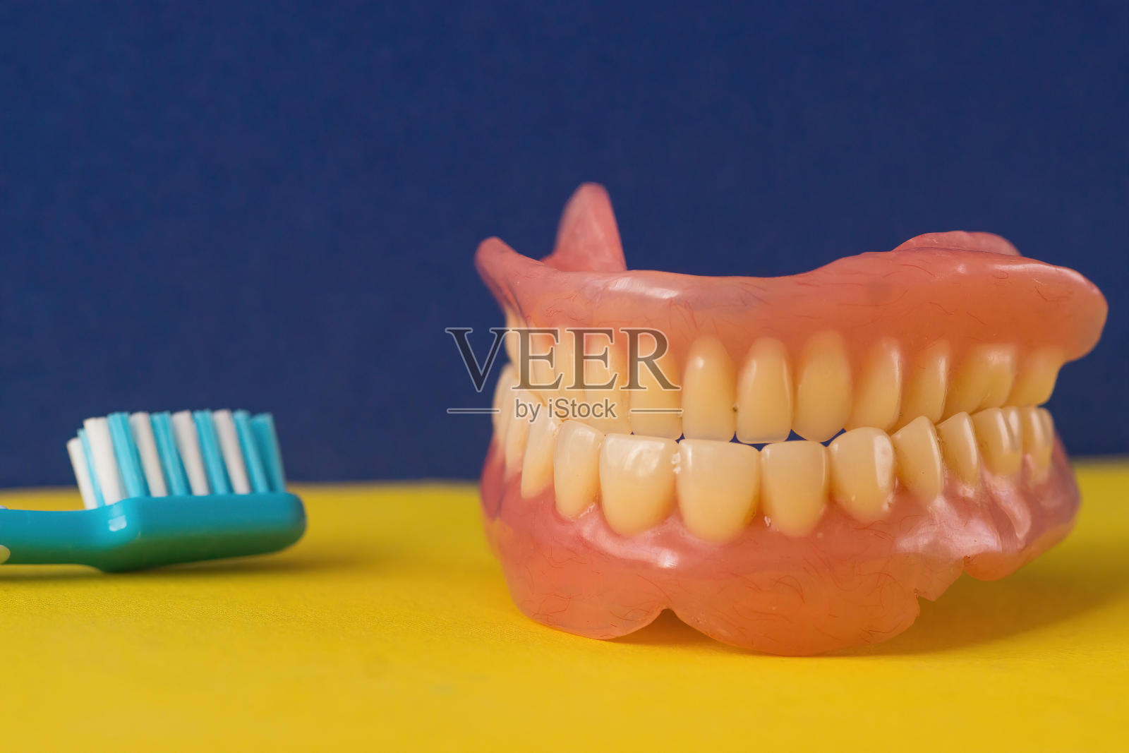 一副人类上颌假牙被放置在亮黄色的表面上，一支蓝色的牙刷水平地放在它的左边。照片摄影图片