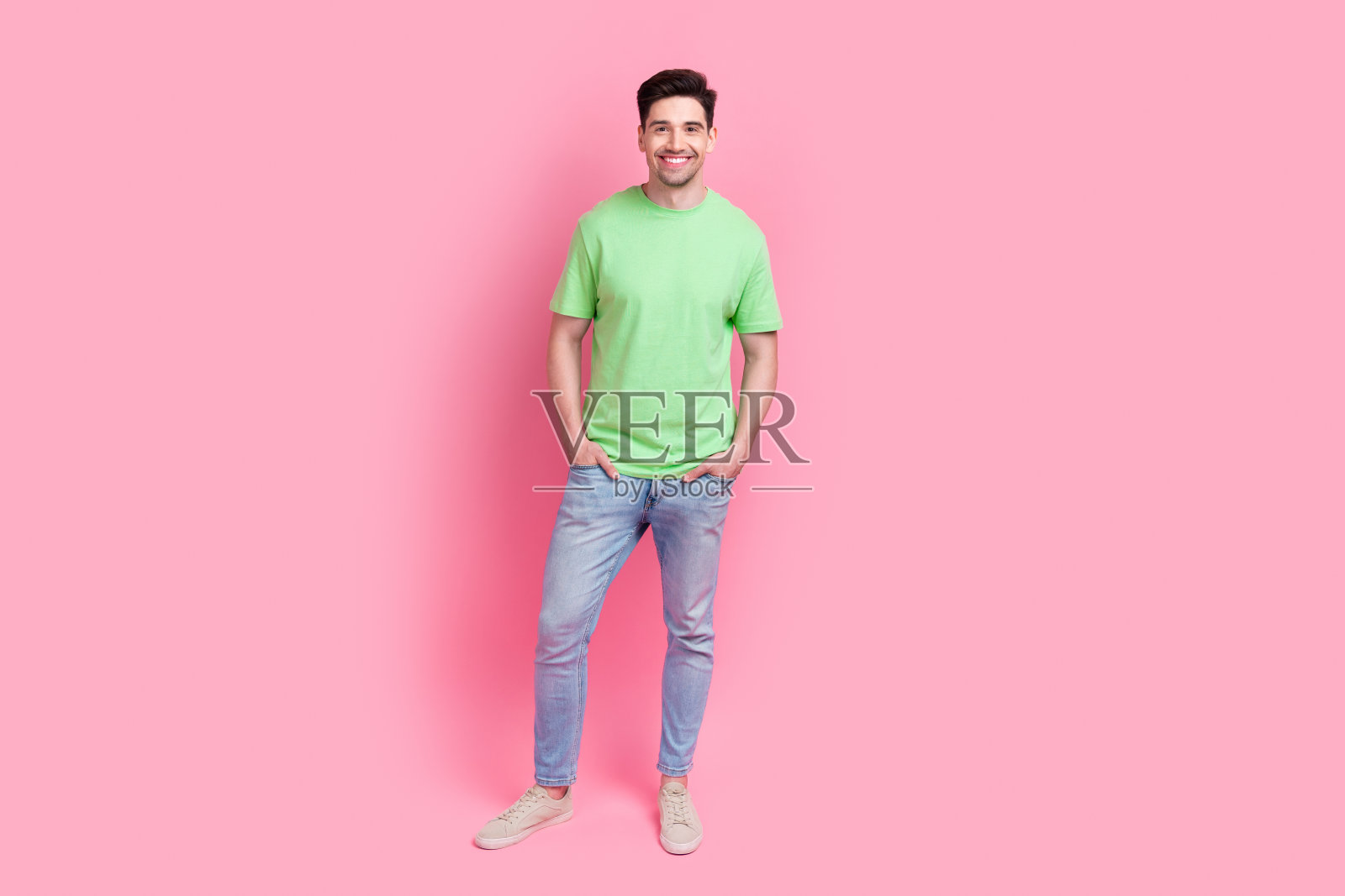 穿着绿色t恤和牛仔裤的年轻帅哥全身照，在粉红色的背景上摆出偷拍的人照片摄影图片