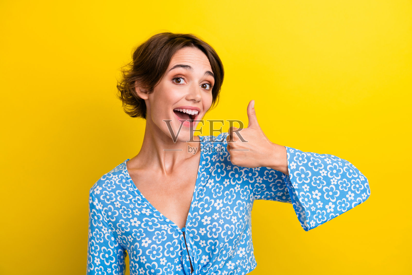 满意的狂喜女孩的肖像，短发，穿着蓝色衬衫，拇指向上，黄色背景上的好作品照片摄影图片