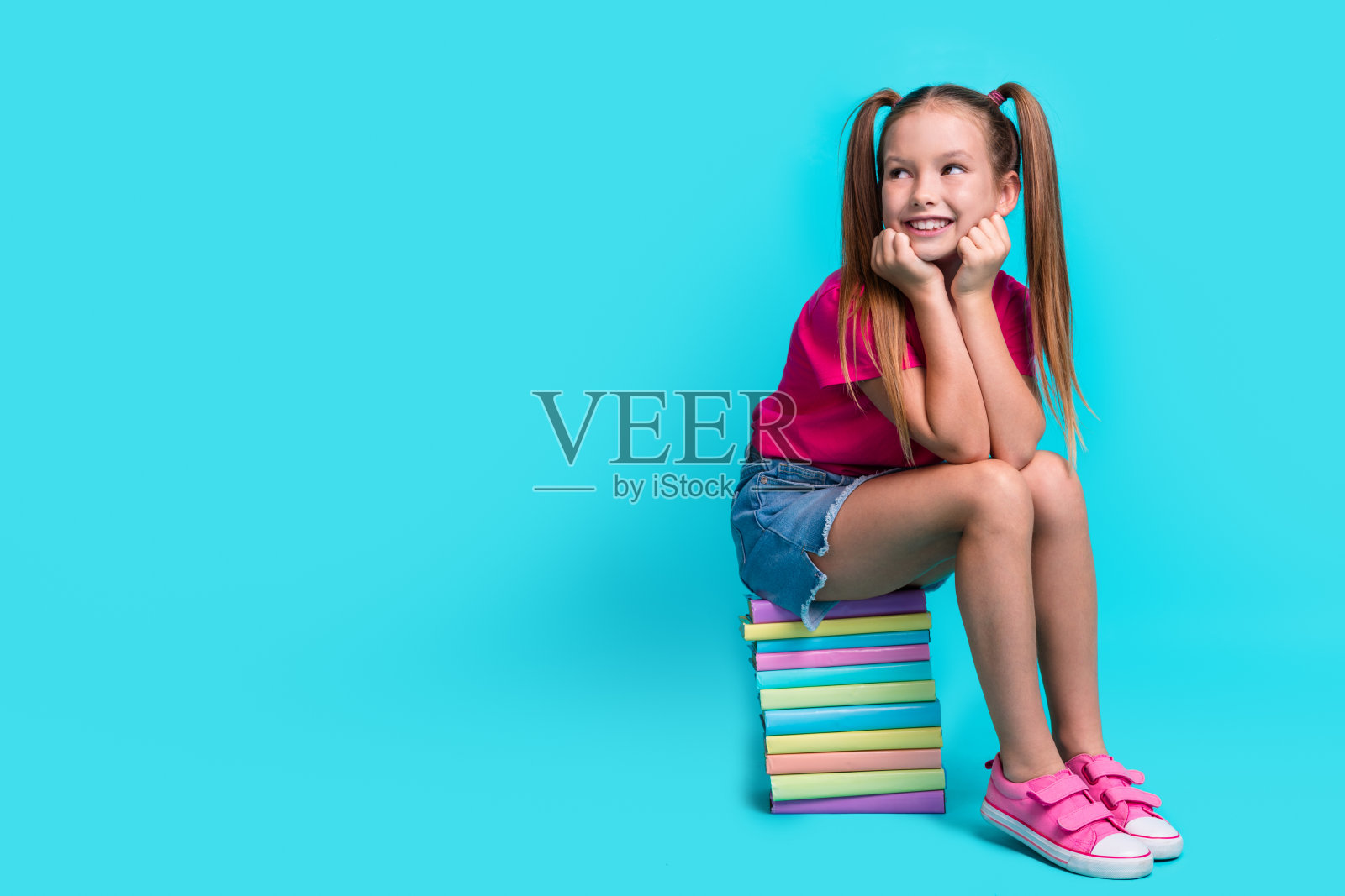 勤奋的女学生穿着t恤的全身照片，坐在书上，看着命题空白的空间孤立在蓝绿色的背景上照片摄影图片