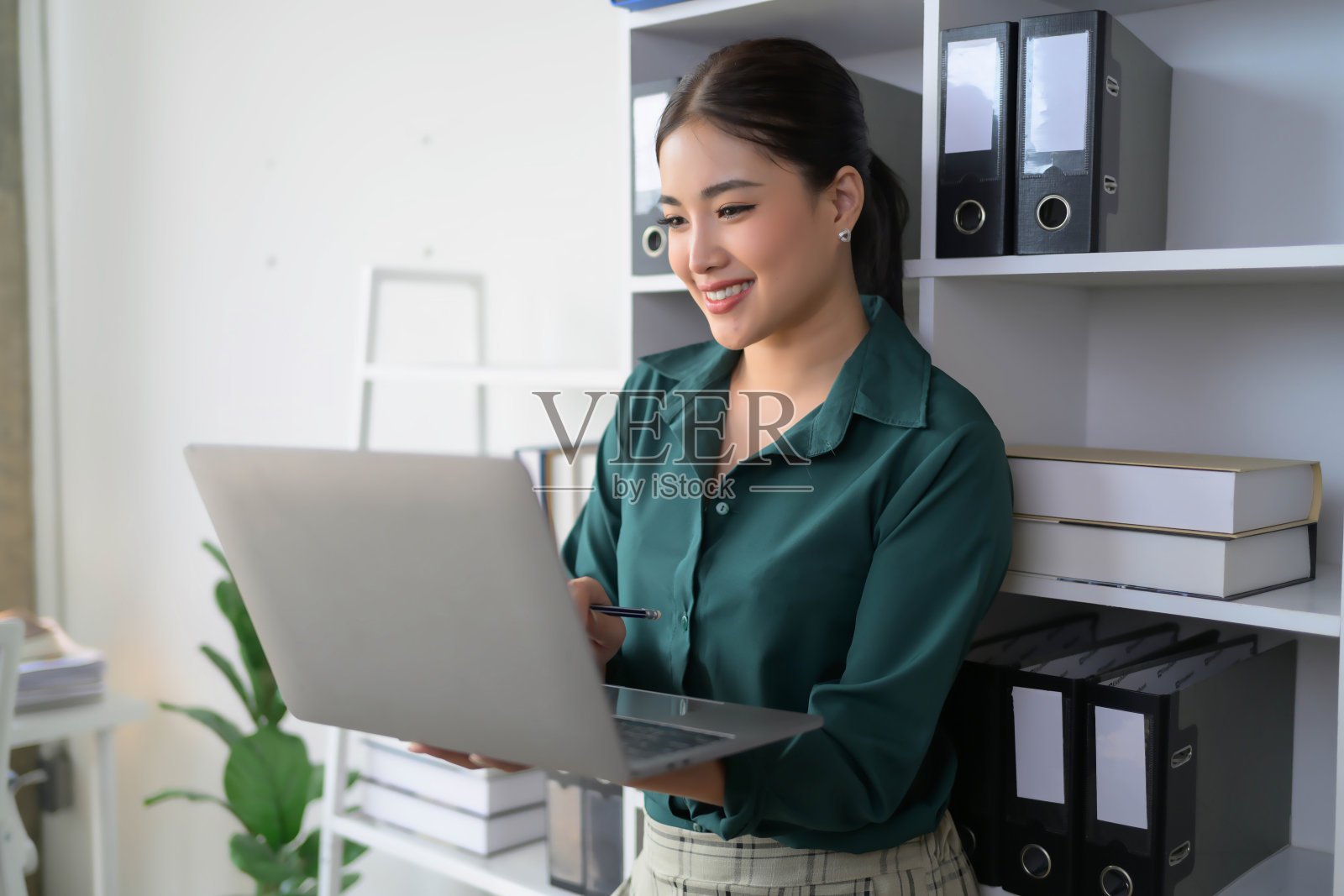 一个女人面带微笑，拿着笔记本电脑站在摆满活页夹的书架前照片摄影图片