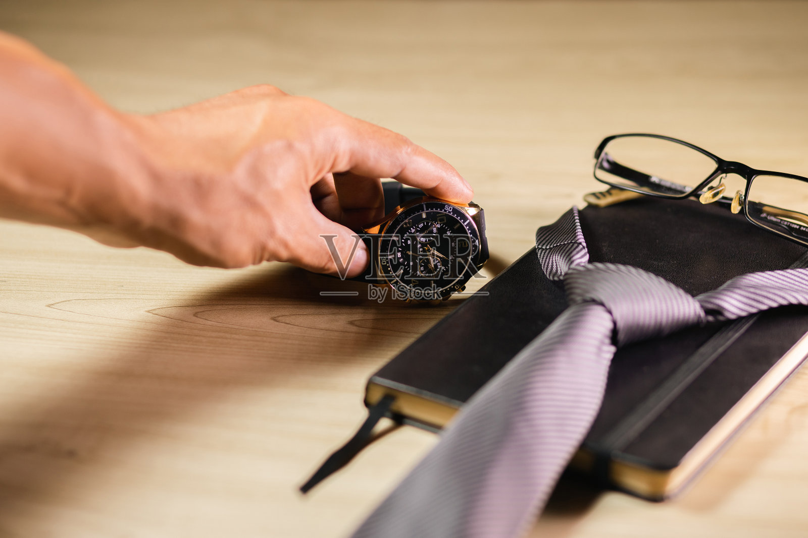 一个男人的手拿起一块手表，旁边是一副眼镜，一个记事本，还有一条放在木架上的领带。父亲节概念。照片摄影图片