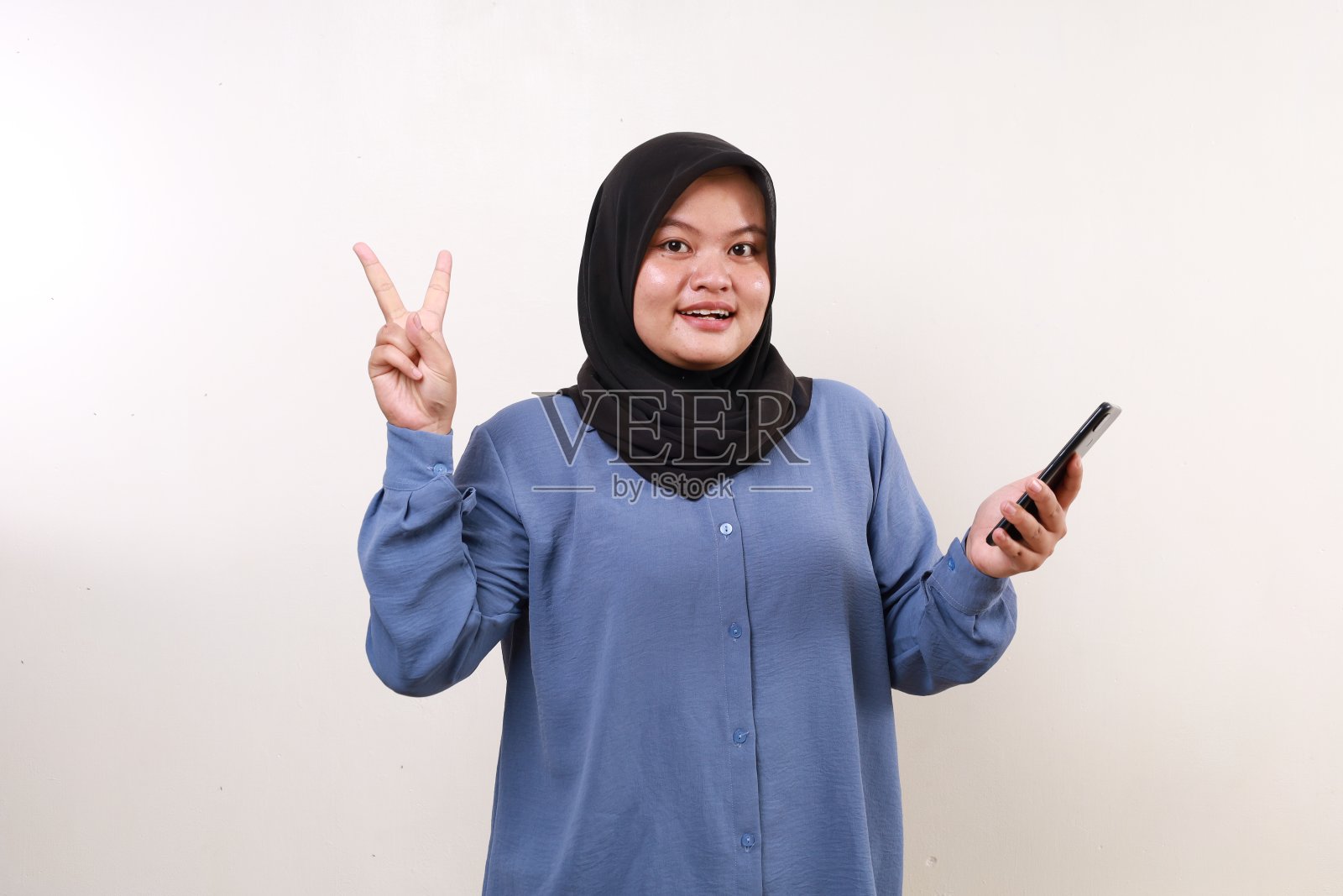 哇，兴奋的亚洲成年女性站在那里，拿着手机，伸出两个手指照片摄影图片