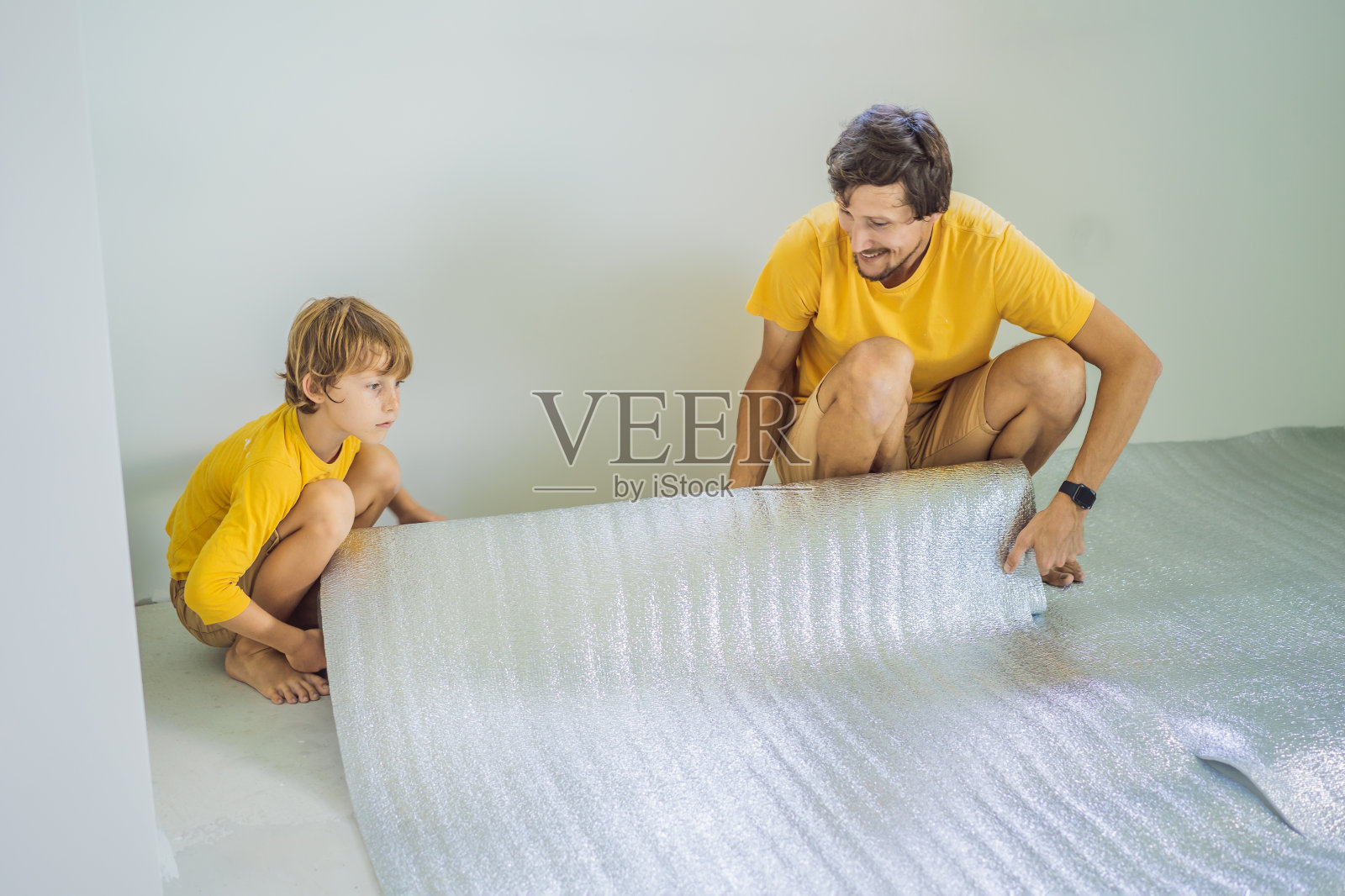 爸爸和儿子正在铺设地板隔热层。卷绝缘布安装一层强化木地板照片摄影图片