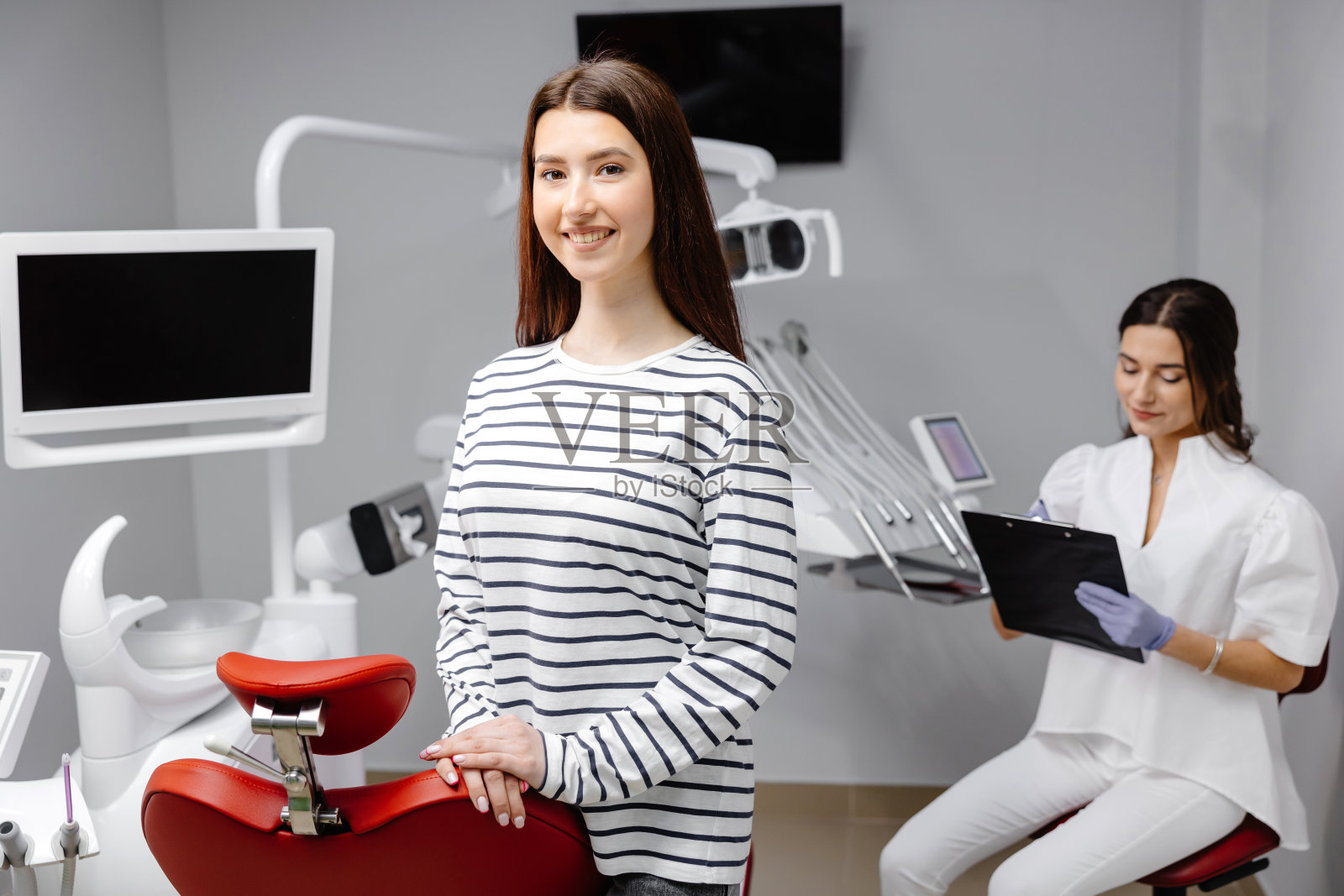 一个年轻漂亮的女孩站在诊所的牙科椅旁，医生正在记录病人的健康状况照片摄影图片