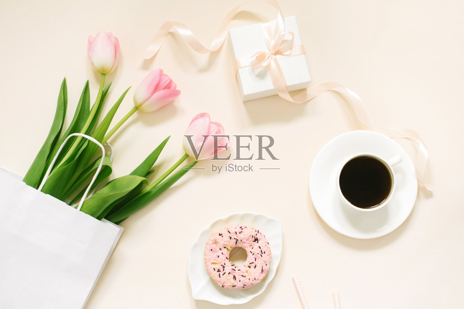 早上一杯咖啡，甜甜圈，礼物或礼品盒和春天的郁金香花在米色的背景。为妇女节、母亲节、情人节准备的美味早餐。平的。照片摄影图片
