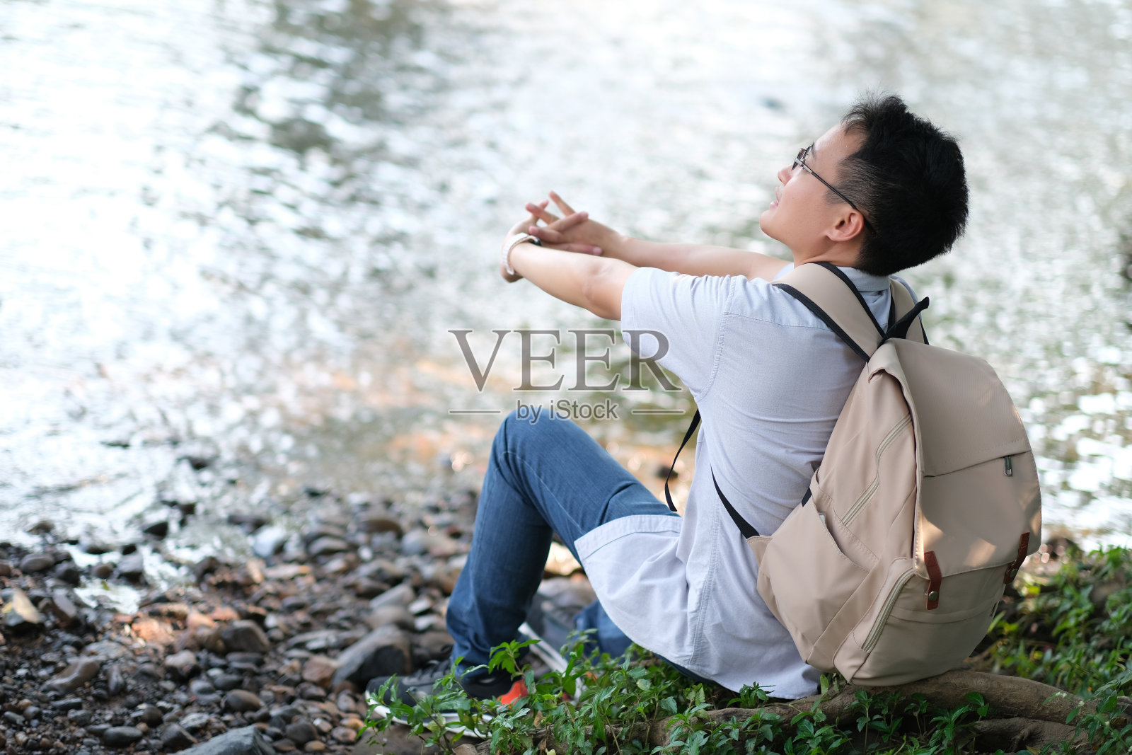 年轻的亚洲人坐在河边的岩石上，抬头仰望，伸展手臂，大自然围绕着他，森林，树木，水带着淡淡的微笑和清新的感觉，人们对自然负责的观念照片摄影图片