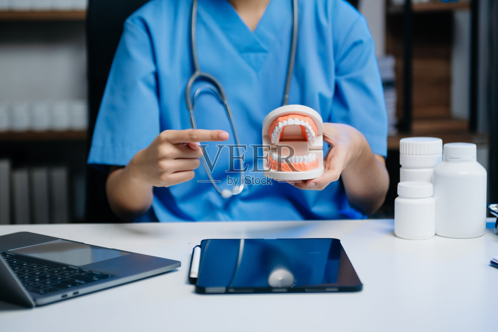在牙科现代化办公室里，牙医坐在桌子旁，拿着颌骨样本和牙齿模型，用平板电脑和笔记本电脑工作照片摄影图片