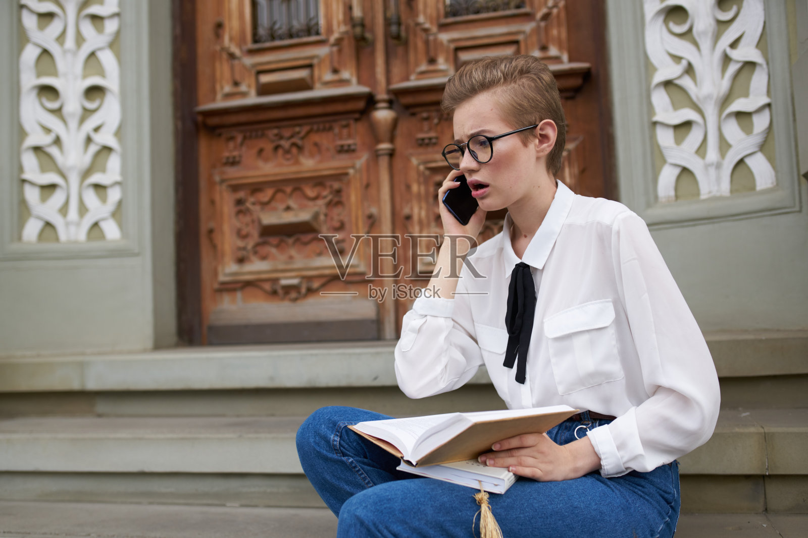 一位女学生坐在大楼附近的台阶上，手里拿着一本书和一部手机照片摄影图片