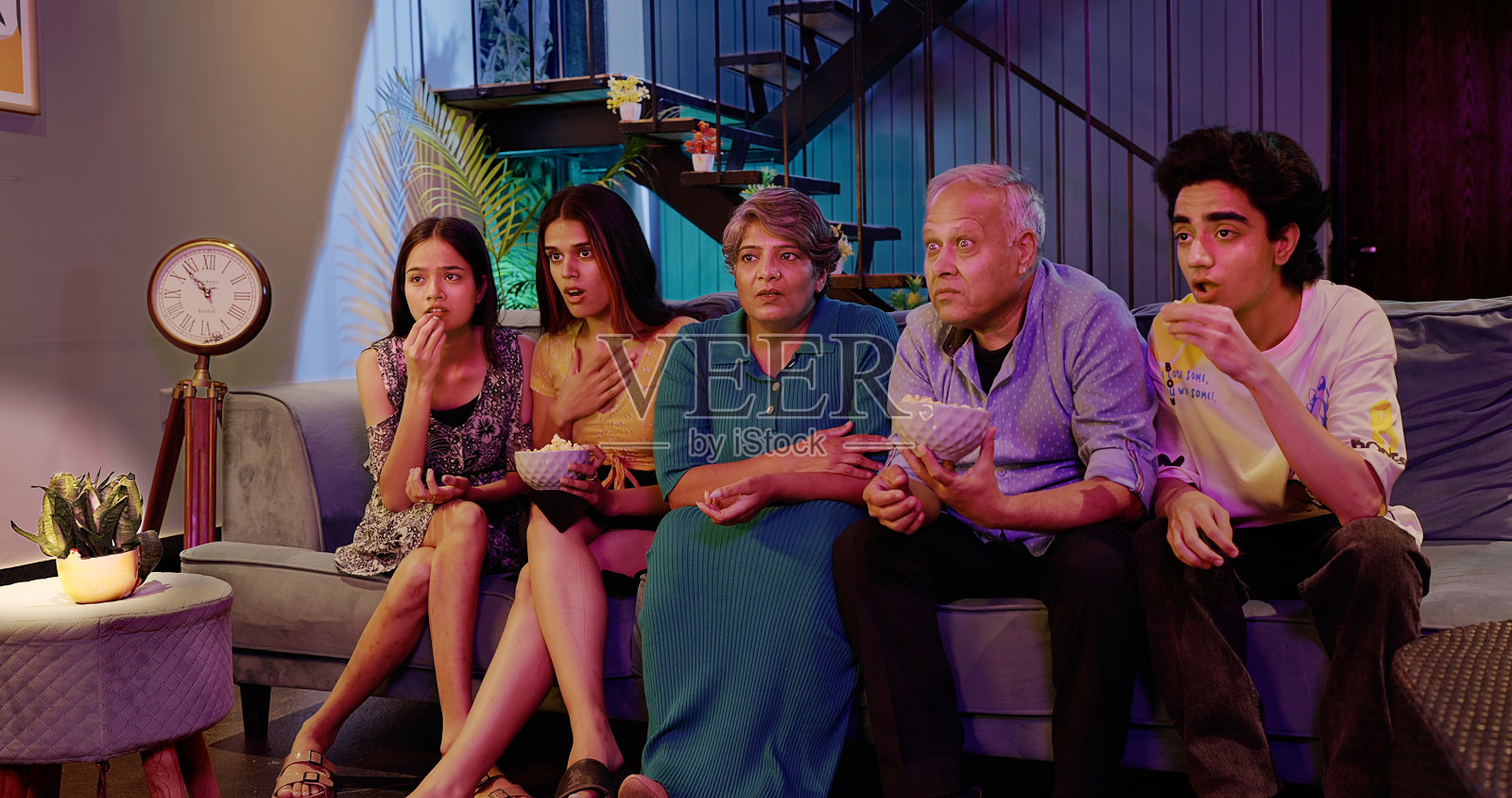 印度幸福延伸的老年家庭坐在沙发上看恐怖惊悚电视，脸上带着恐惧的表情，年长的祖父母，年轻的十几岁的男孩，女孩，拿着碗，一起吃零食爆米花，深夜在家里看电影照片摄影图片