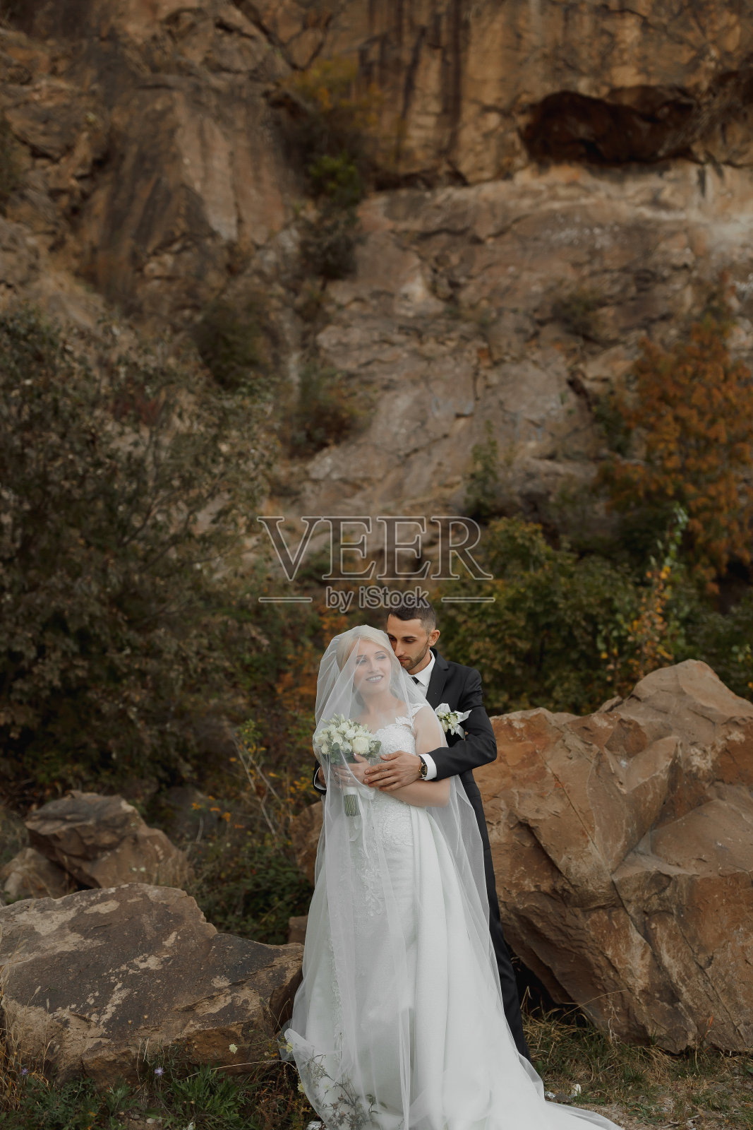 新娘和新郎站在高山上的一块岩石上，旁边是一块大石头。新娘戴着面纱，新郎拥抱着她的肩膀照片摄影图片