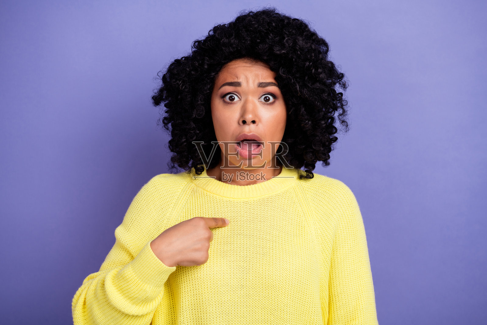 被冒犯了的吃惊的女人，留着非洲式发型，穿着黄色毛衣，在紫色的背景上看着自己照片摄影图片