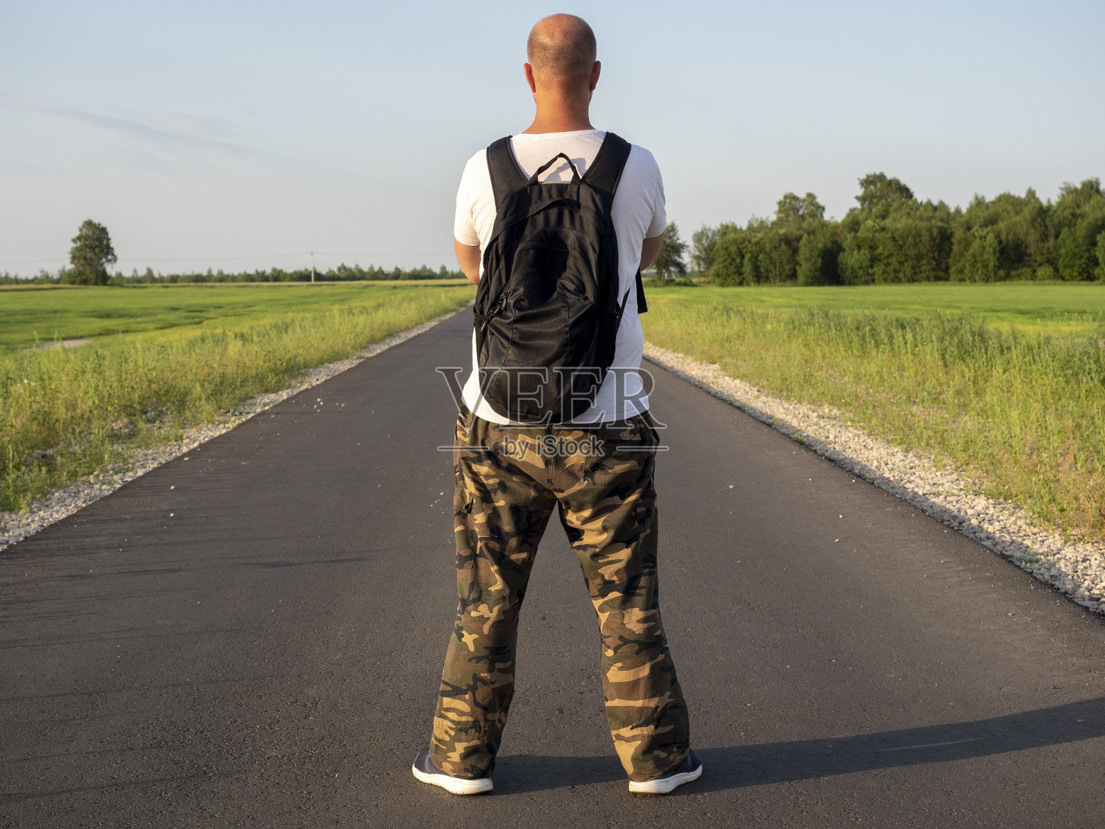 一个背着黑色背包的中年男子站在柏油路上，背对着镜头。那人双臂交叉在胸前。旅行和新发现的概念照片摄影图片