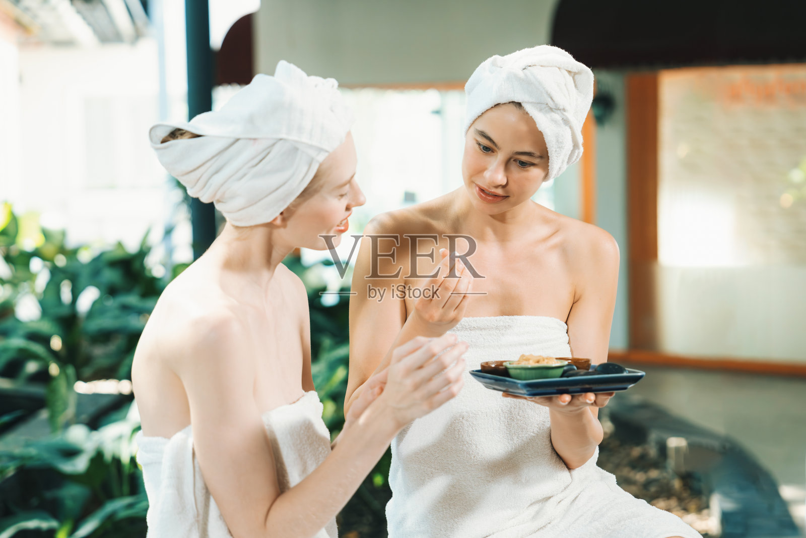 两个漂亮的女人裹着毛巾，一边拿着草药碗一边咯咯地笑。宁静。照片摄影图片