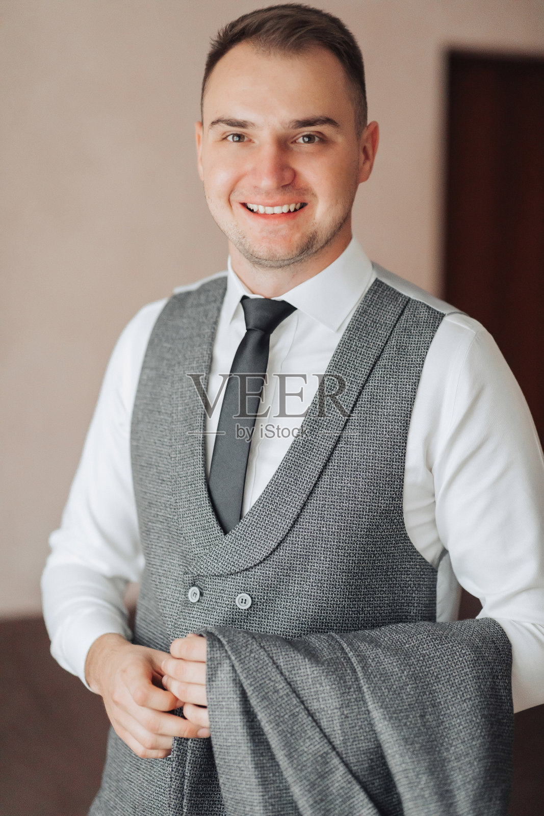 一名身穿白色衬衫和灰色背心的男子拿着夹克，站在窗前看着镜头。前视图。一块时髦的手表。男人的风格。时尚。业务照片摄影图片