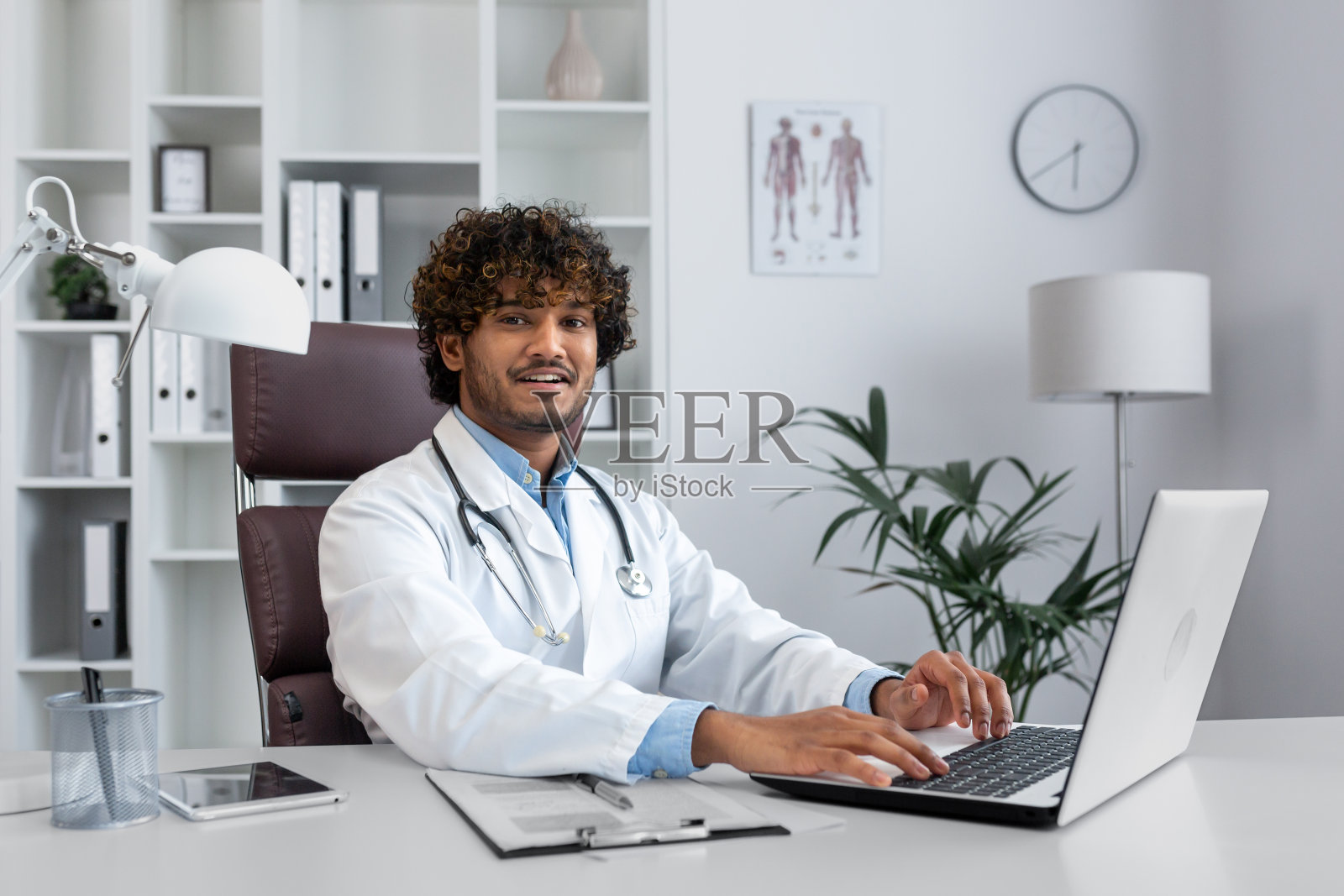 一位年轻的西班牙裔医生在办公室里用笔记本电脑工作，穿着白大褂，戴着听诊器坐在桌子旁，微笑着看着镜头照片摄影图片