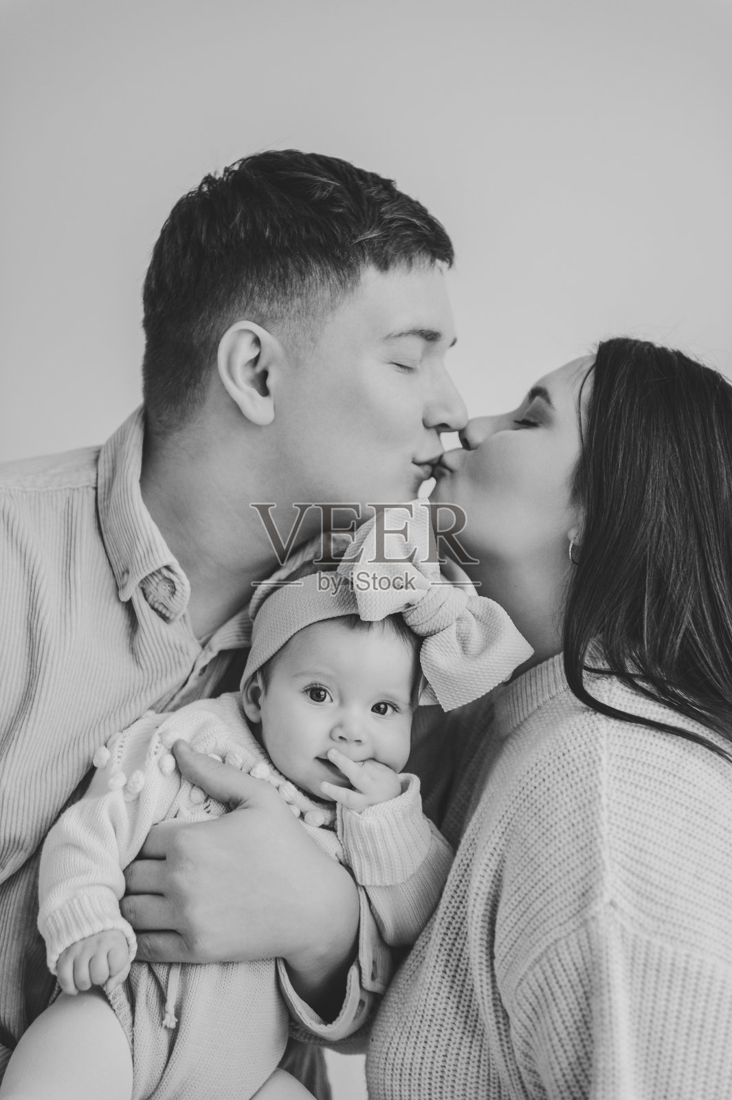 父亲亲吻母亲拥抱女儿。幸福的家庭在节日里拥抱可爱的宝宝。爸爸，妈妈牵着婴儿的手。爸爸，妈妈抱着小女孩，头发上扎着蝴蝶结，孤立在墙上。黑白照片。家庭。照片摄影图片