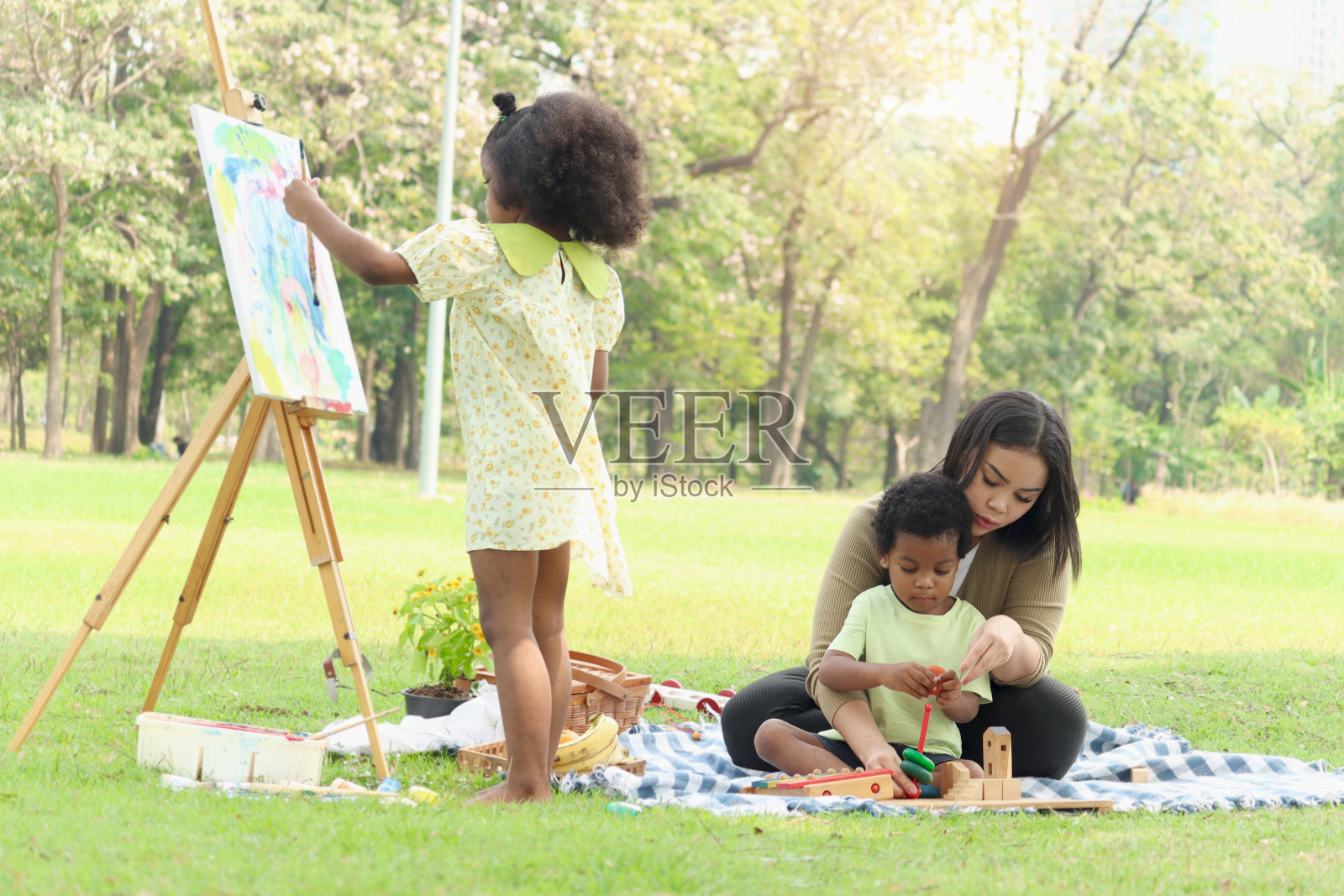 可爱的微笑，黑色卷发的亚非女孩在画布上画画，母亲和弟弟在户外花园玩耍，孩子和父母一起在夏日公园野餐，幸福的家庭。照片摄影图片