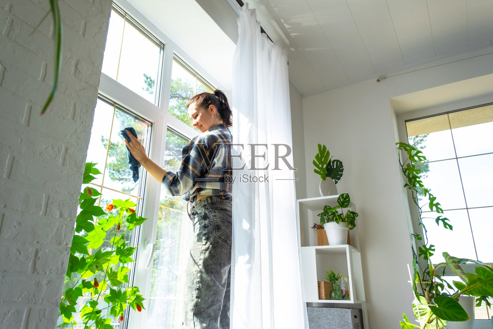 妇女用带有喷雾清洁剂的抹布和拖把手动清洗房子的窗户，室内有白色窗帘。恢复春天的秩序和清洁，清洁服务照片摄影图片