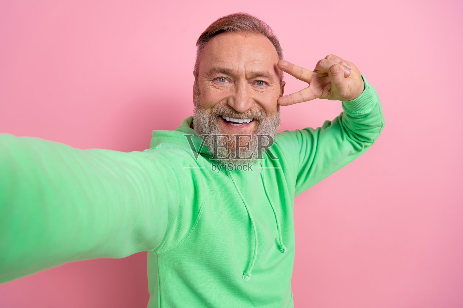心情好时髦的家伙穿着绿色卫衣，带着v字手势，自拍孤立的粉红色背景照片摄影图片